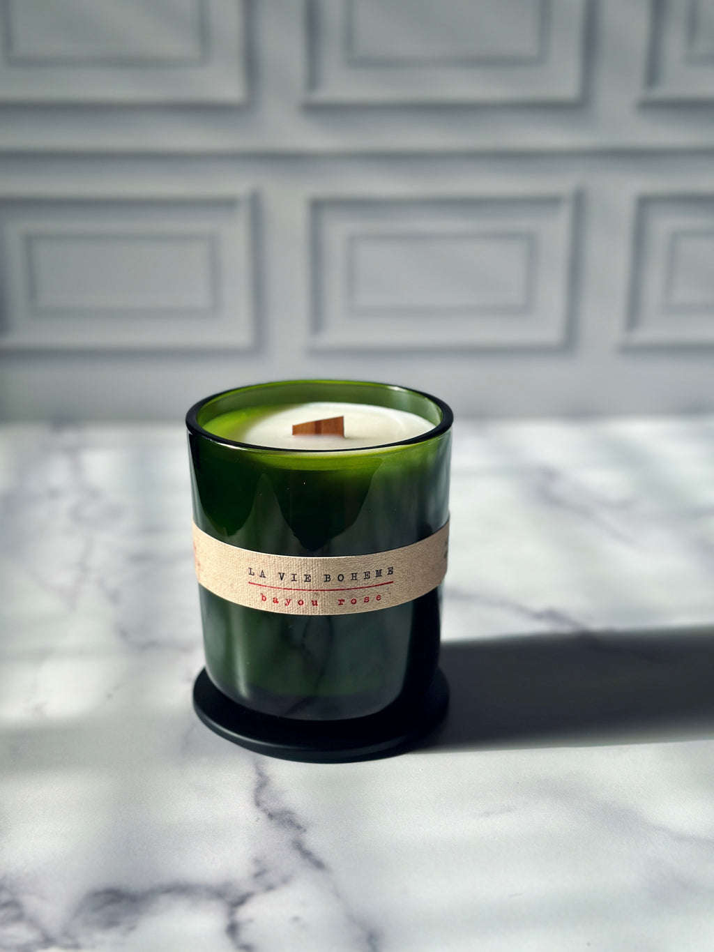 Green glass jar candle with black lid, typewritten label, wooden wick, on a marble background